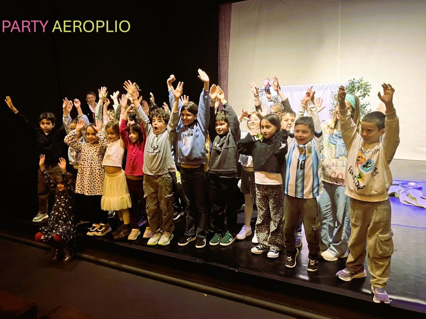 Children in a line on stage with their arms raised, at the end of the party